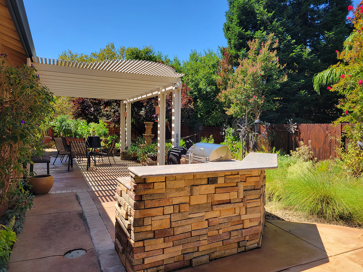 Mary Kay Williams backyard garden showing a paved and shaded patio area with a grill and chairs surrounded by lush green plants and bright red flowers.