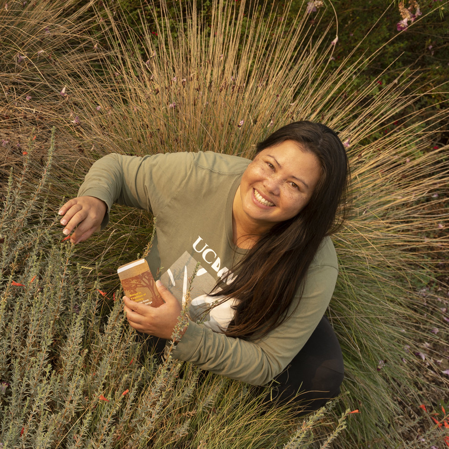 Image of woman with long dark straight hair looking up into the camera while she spreads seeds in a verdant garden.