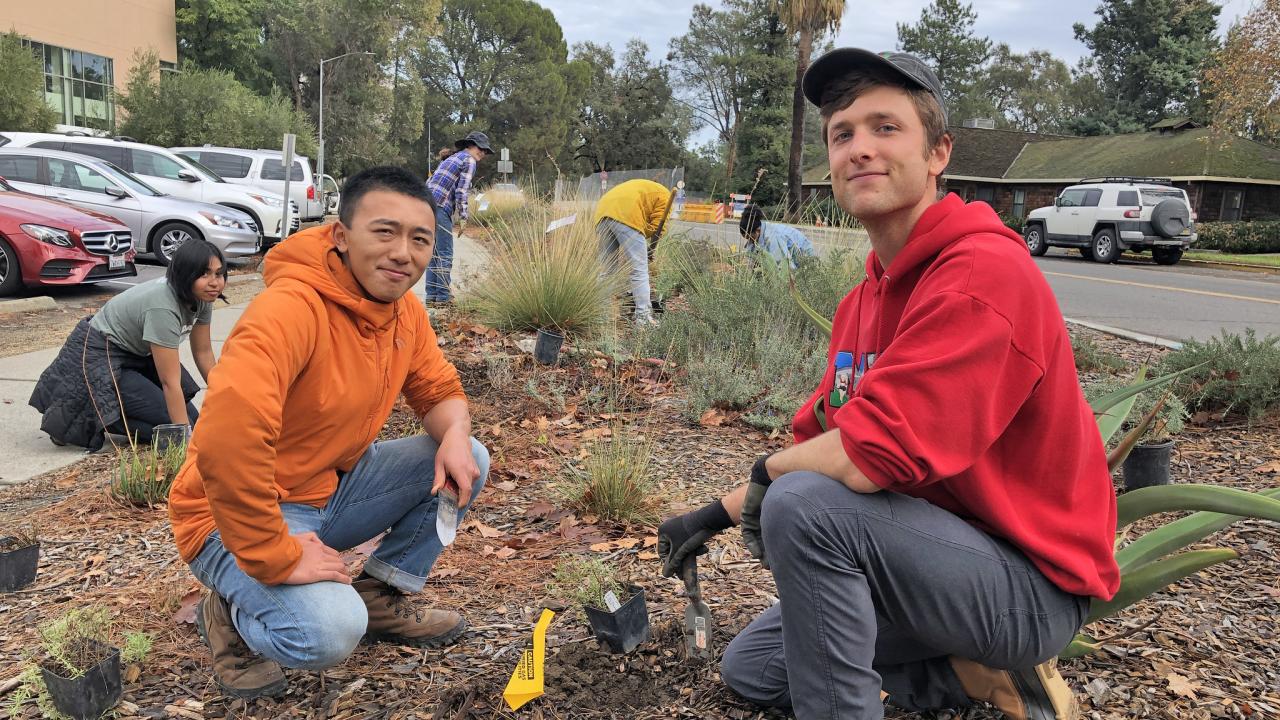 Program co-coordinators Chris Huang and Cameron Long take a break from planting a new plant to pose for a photo, kneeling with the landscape in the background.