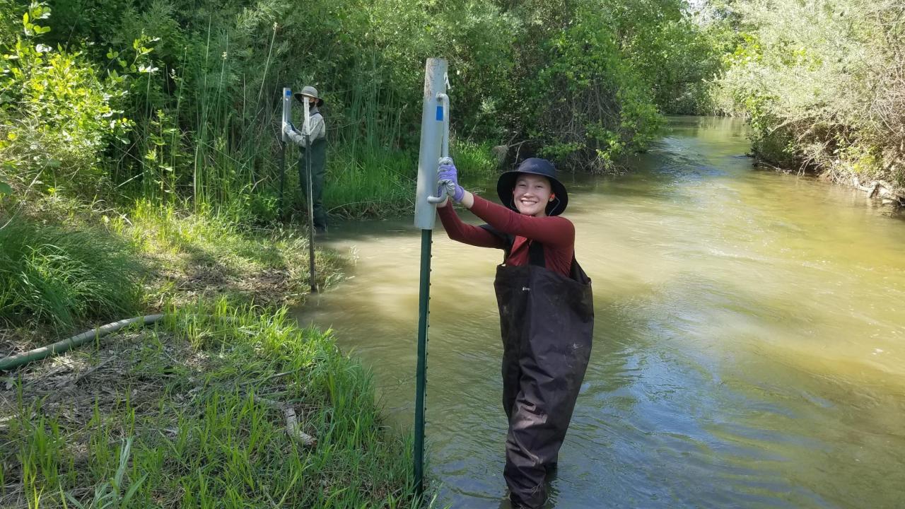 Two student interns pose smiling, wearing waders standing in Putah Creek